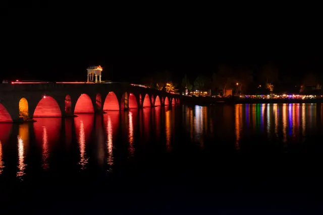 A stone arched bridge lit up at night, reflected in the Tunca River