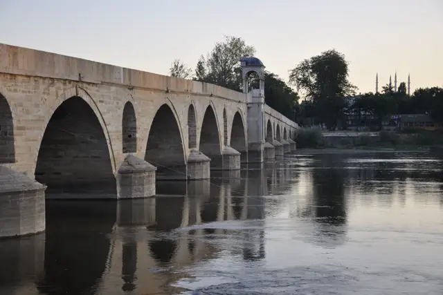 The arches of the Meriç Bridge at sunset, reflected in the calm river, with the Selimiye's minarets in the distance